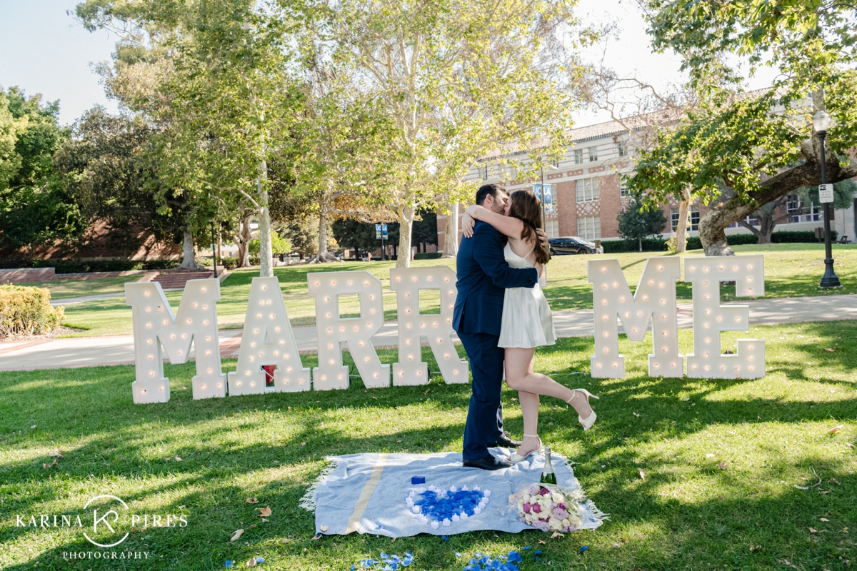 Man on one knee proposing to his fiance in front of a ‘Marry Me’ sign at UCLA