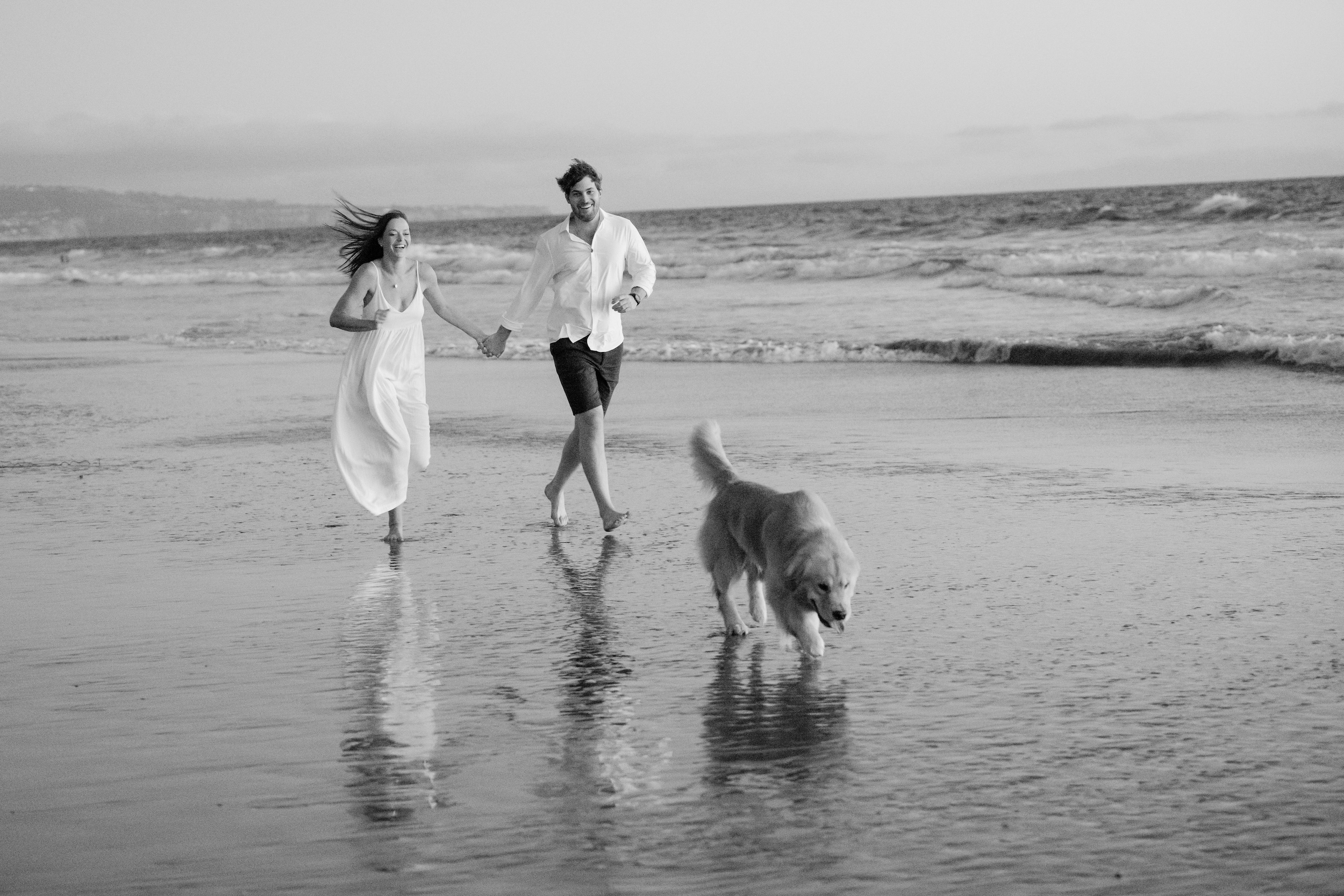 A couple walking barefoot along the shoreline with golden retriever Gundo trotting beside them at Manhattan Beach, for their engagement session.